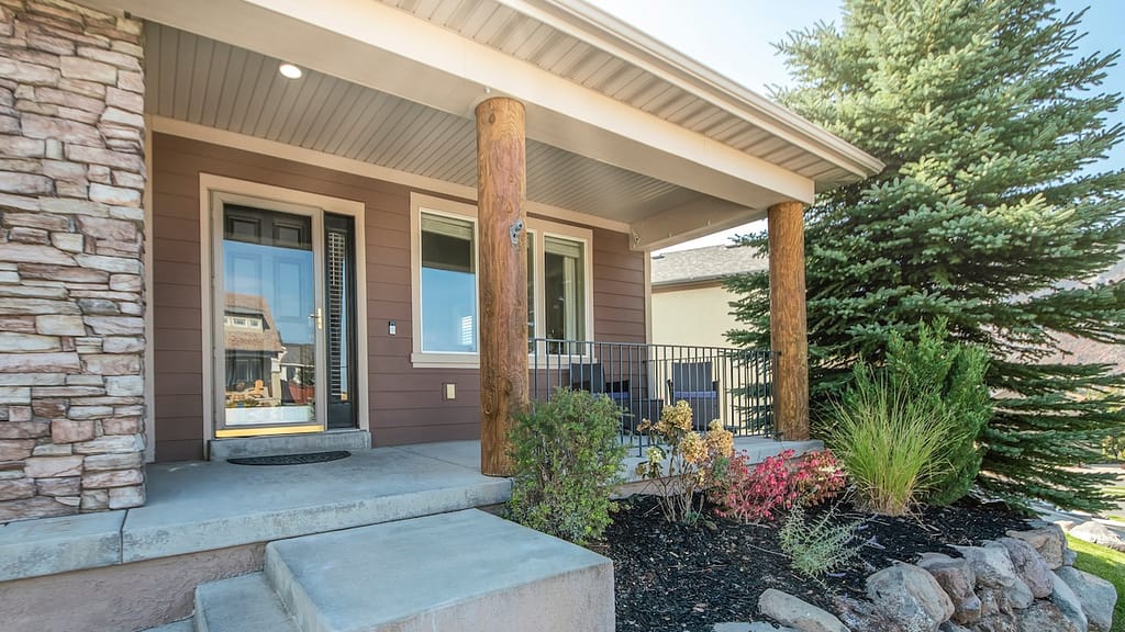 Pano Front exterior of a house with a raised edge landscape and stone brick walls. A porch with storm door, windows and log posts with a steel railings, and a concrete stairs near the garage with bricks.