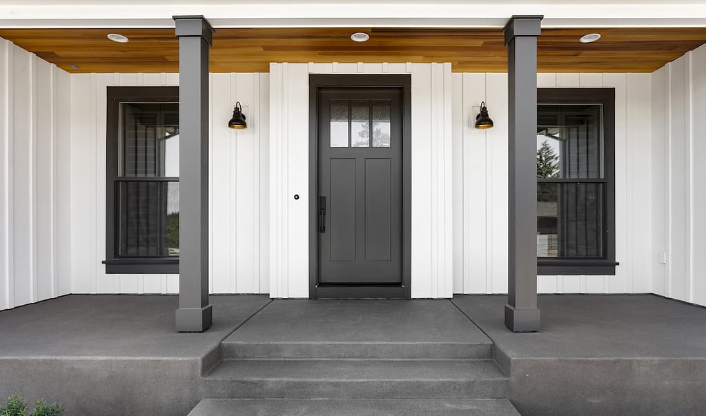 Front door to modern farmhouse home. Home exterior with white vertical wood siding and black front door. Columns and sconce lights on both sides of door.