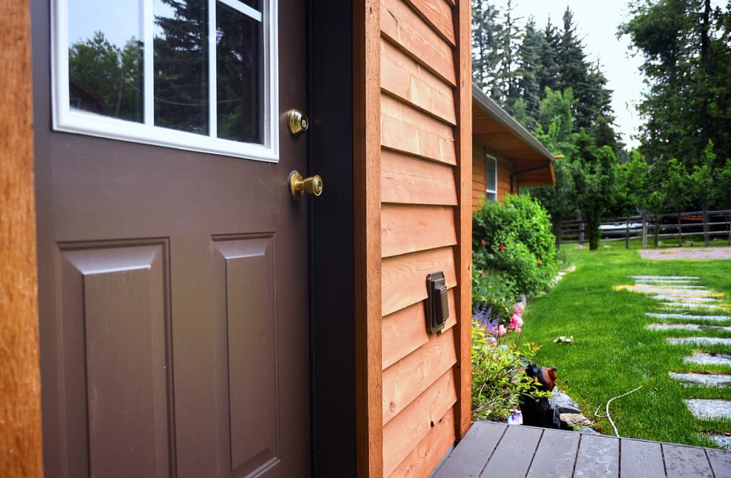 A wooden house entrance features a brown door and cozy porch, complemented by a stepping stone pathway through the lush green lawn. Trees and shrubs thrive under an overcast sky, enhancing the rustic charm. The clean wood siding adds to the welcoming allure of this serene retreat.