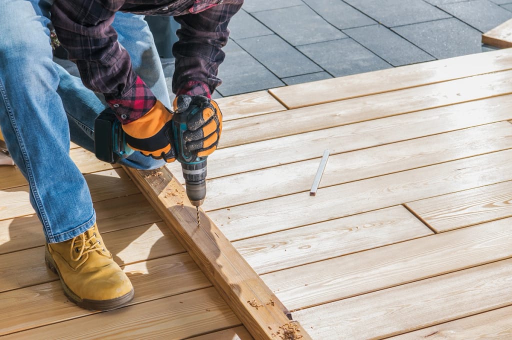 A person wearing gloves and boots is busy with deck repair, using a drill to secure a wooden plank. Kneeling amid an array of tools and planks, they work on the newly constructed surface of light-colored wood.