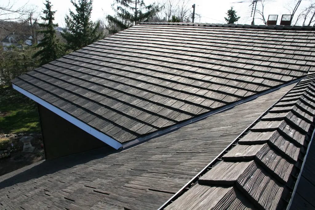 A roof with dark, overlapping rubber shingles. The roof is sloped and extends into the distance. Surrounding trees and a partly cloudy sky are visible in the background.