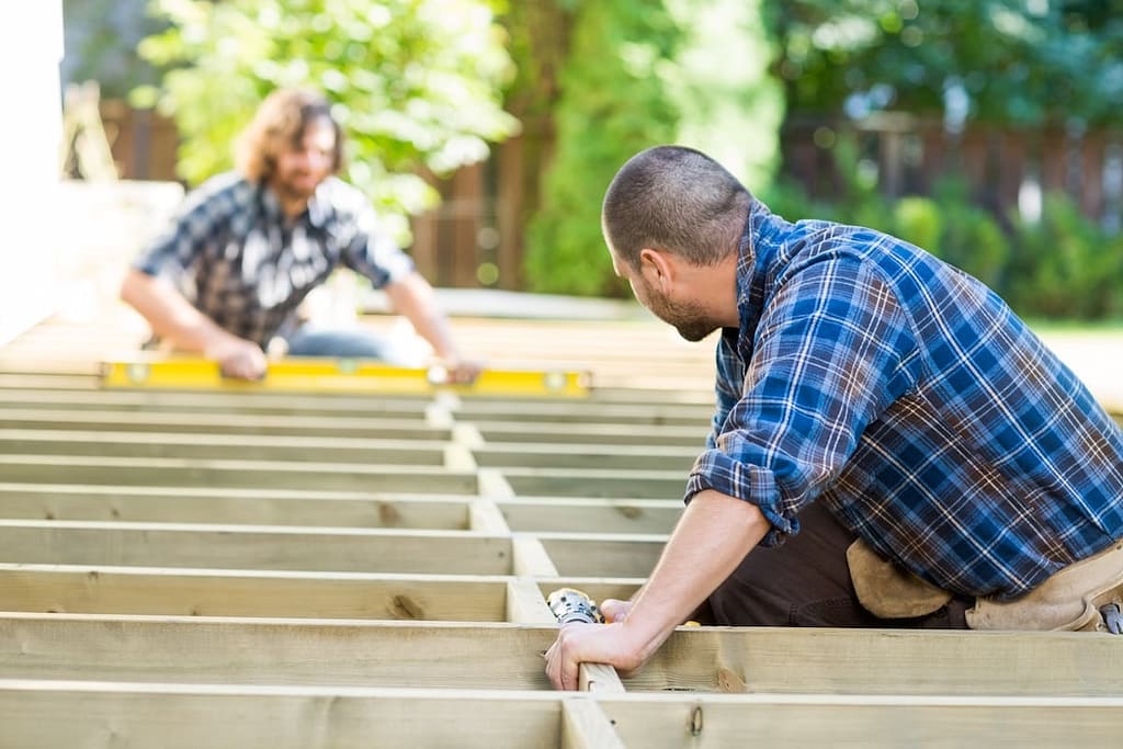 Two men in plaid shirts are building a wooden deck frame outside