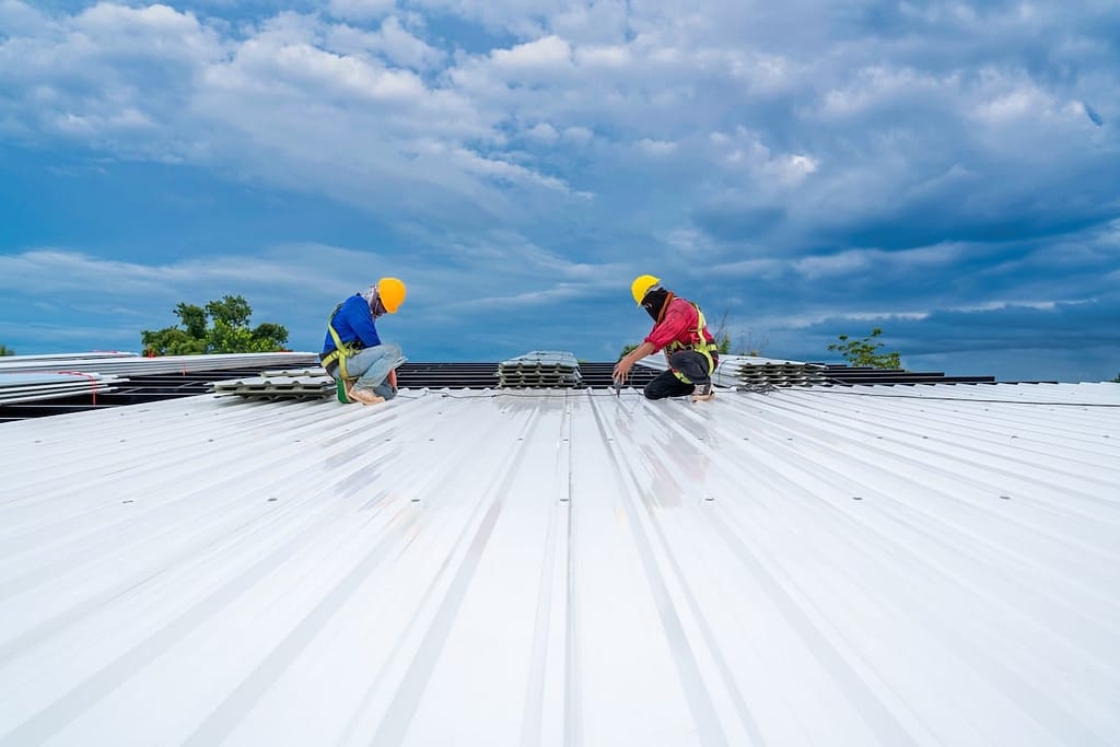 Two construction workers in yellow helmets are installing metal roofing sheets on a bright white roof