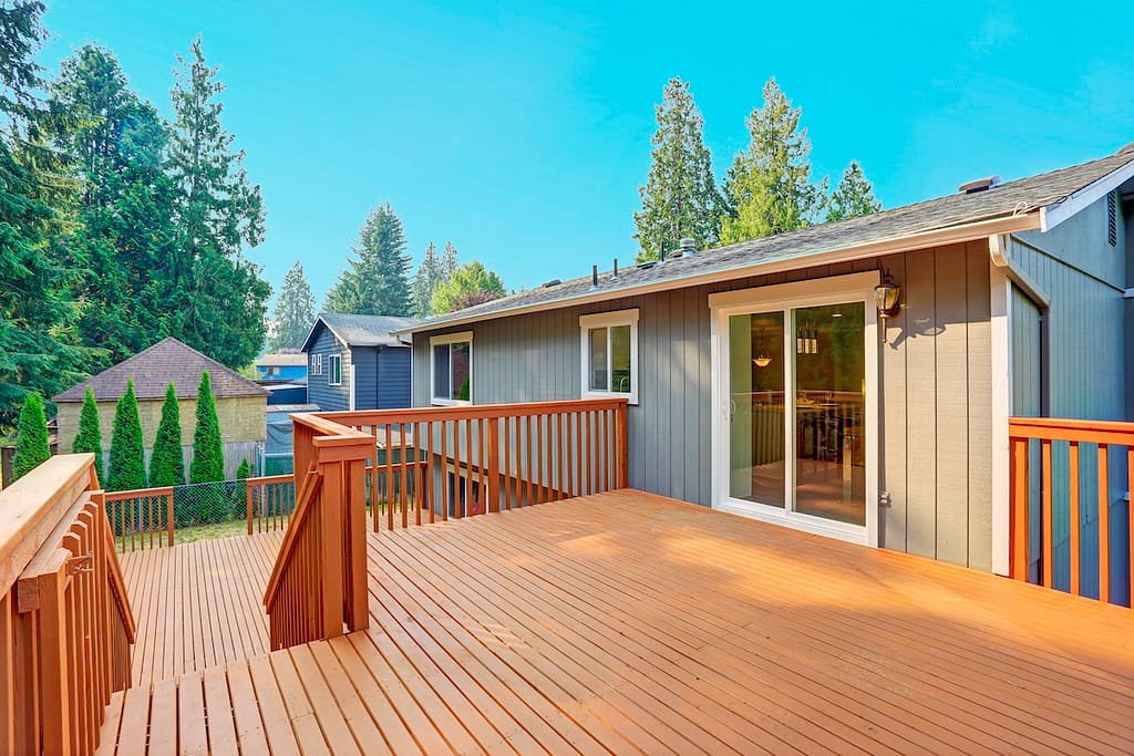 A spacious wooden deck extends from a gray house with sliding glass doors, surrounded by trees. The sky is clear and blue, creating a serene outdoor setting. Other houses are visible in the background.