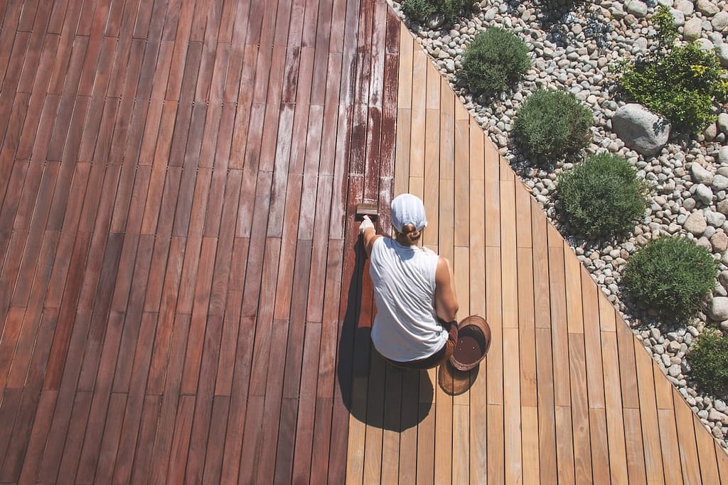 Person painting a wooden deck in two shades of brown, with neatly arranged green shrubs and gray stones on one side.