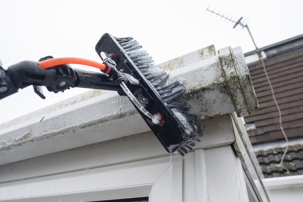 A close-up of a brush attached to a pole being used to clean dirt and mold from the white guttering of a house