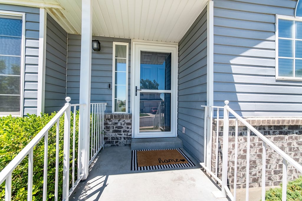 Front porch of a house with glass storm door with sidelight. Entrance of a house with doorbell and doormat at the doorsteps with railings and an exterior of a gray vinyl lap and bricks sidings.