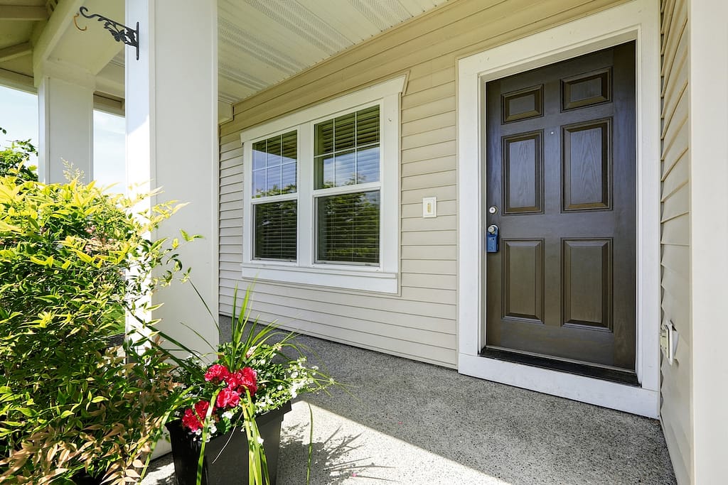 Open porch with concrete floor, column and entrance brown door. Decorated with hanging lantern light and pots with flowers