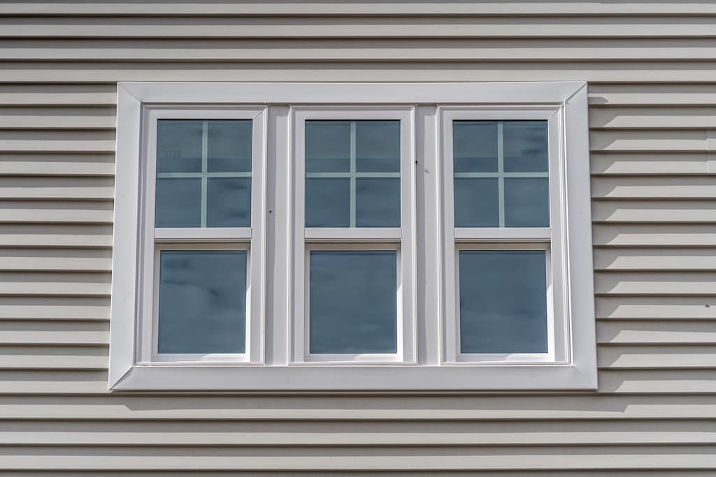 Triple hung window with fixed top sash and bottom sash that slides up, sash divided by white grilles a surrounded by white elegant frame horizontal white vinyl siding on a new construction residence