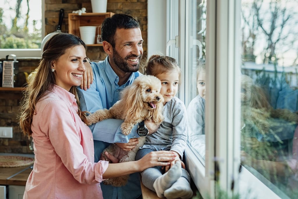 Young happy family and their dg relaxing by the window at home and looking through it.