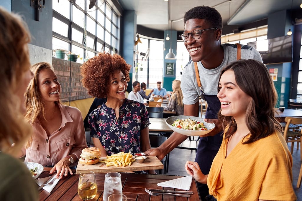 Waiter Serving Group Of Friends Meeting For Drinks And Food In Restaurant