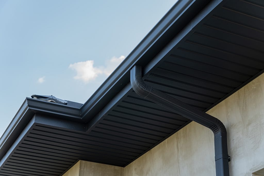 Close view of a roof edge and downspout against a bright blue sky highlights contemporary construction features and materials.