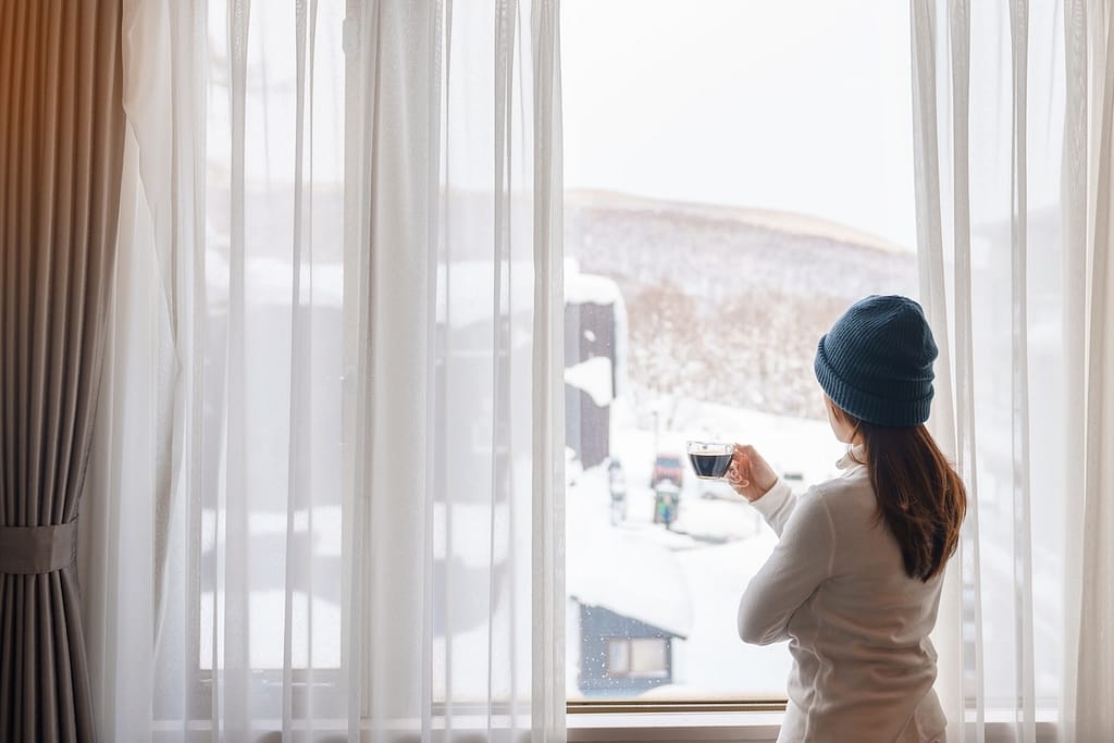 young woman in sweater with cup of coffee looking through the window in winter season, happy female enjoying snowfall outdoor view at apartment or home in the morning. Waking and Relaxing concept