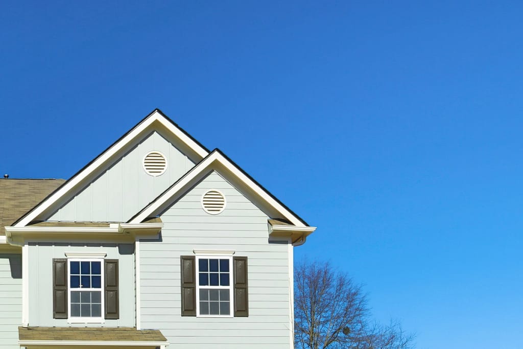 The corner of a light gray house with two small peaked roofs stands out against the clear blue sky, showcasing well-maintained siding. It features two windows with dark shutters, and a tree with bare branches is partially seen in the background.