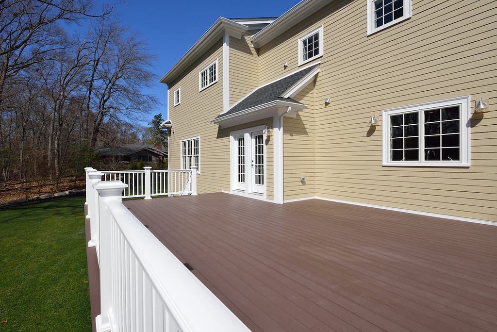 A large composite deck extends from a beige house with white trim and multiple windows. The deck features white railings and overlooks a grassy yard with a backdrop of leafless trees under a clear blue sky.