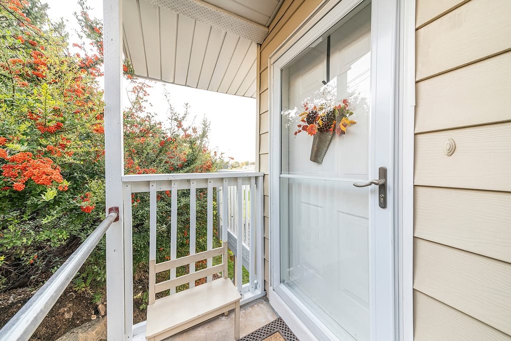 Entrance of a house with glass storm door over the white door with hanging flower decoration. Front porch of a house with railings and wooden chair against the shrubs on the background.