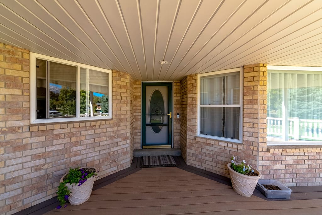 House porch with bricks and potted plants at the front door with oval panel behind the screen door. There is a bay window on the right and sliding window on the left with reflective glass panes.