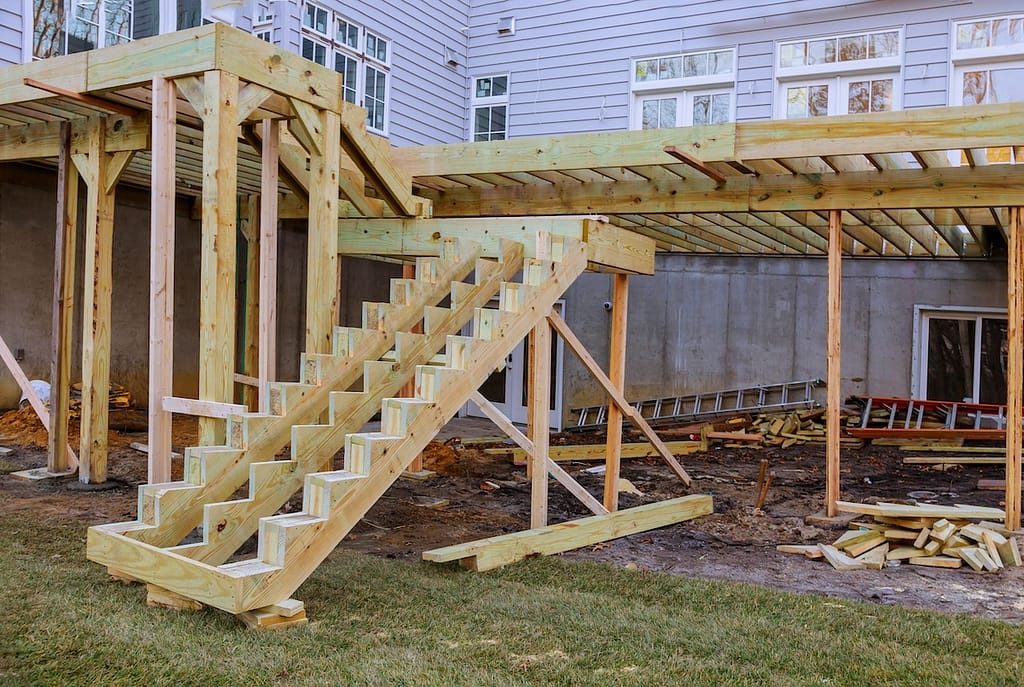 Partially constructed wooden deck and stairs attached to a house, with building materials and tools scattered on the ground below.