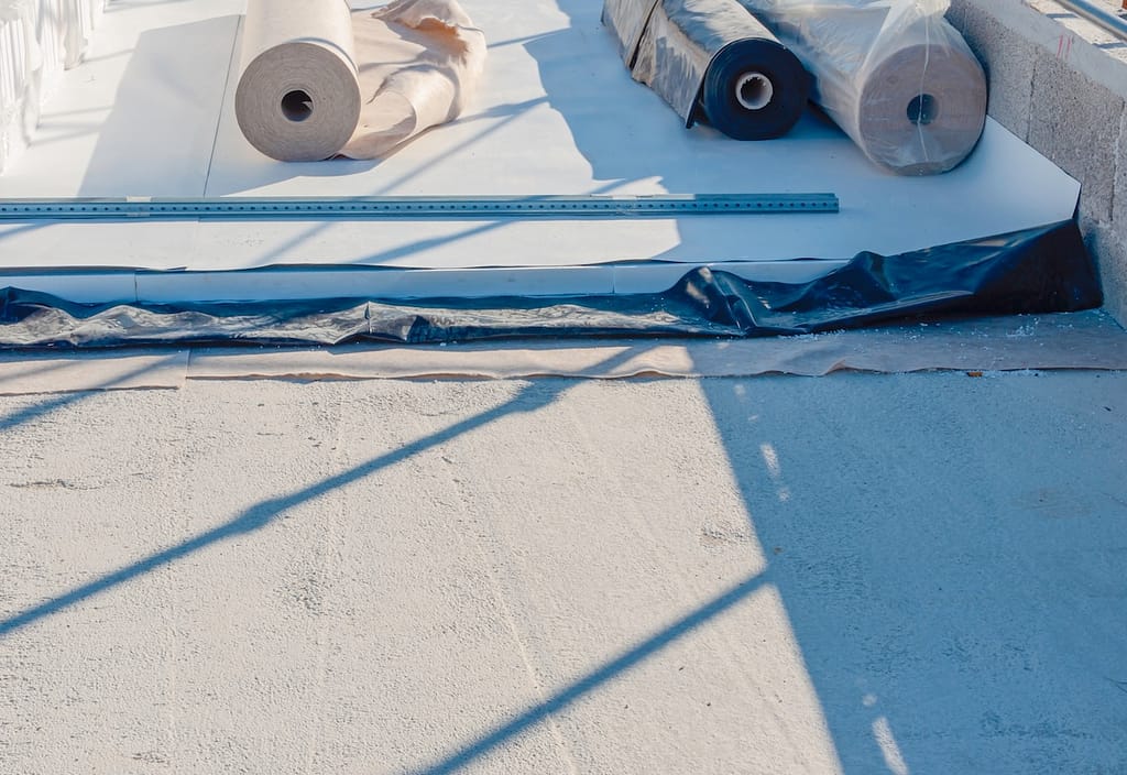 Rolls of TPO roofing material and a metal straightedge lie on a flat, unfinished rooftop.