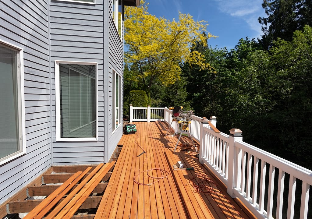 A partially completed wooden deck is attached to a gray house with white trim. Construction tools, lumber, and materials are scattered on the deck