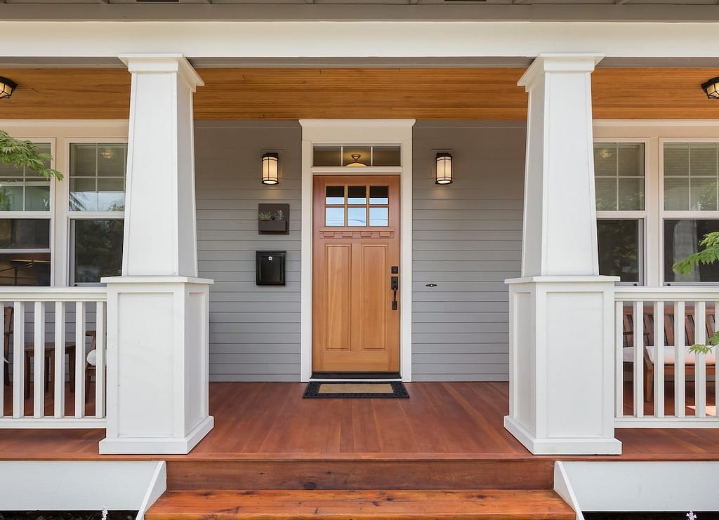 A welcoming front porch with a wooden door centered between two white columns. The house has gray siding and three lit wall lanterns.