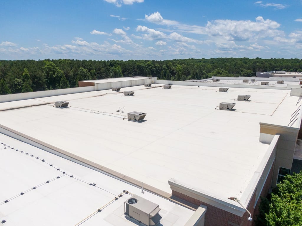 A large, flat white rooftop with several HVAC units is seen against a backdrop of a green forest and a partly cloudy blue sky.