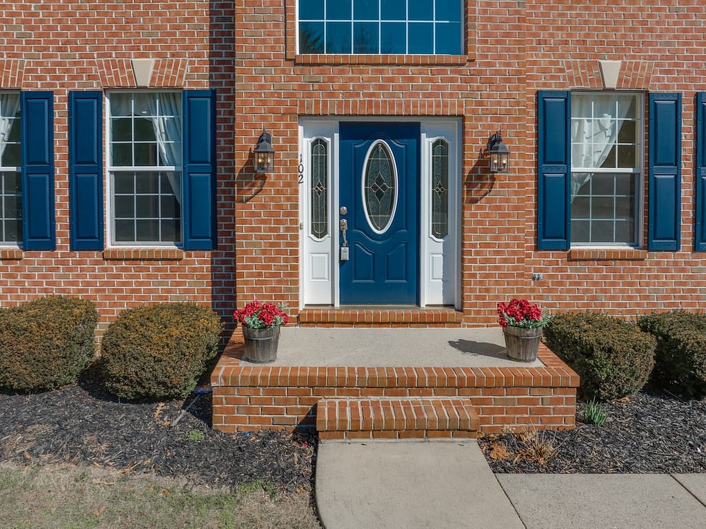 Close up view of elegant blue wooden door with oval stained window, with white sidelights, silver brass color door knob, iron wall light on red brick facade, red flower pot front door colors