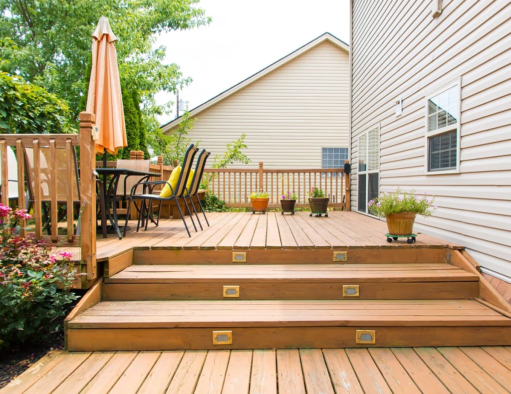 A wooden deck with steps leading to a patio area, featuring a table with chairs and an orange umbrella. Potted plants are placed on the railing and steps. Two houses and greenery are visible in the background.
