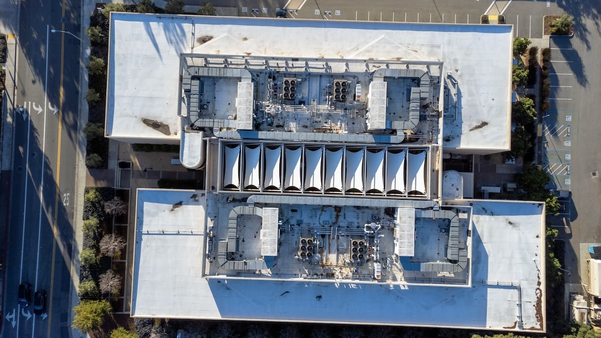 Aerial view of a large, rectangular industrial building with a white roof. The roof features two sections of ventilation or HVAC systems and a central row of triangular, tent-like structures. Surroundings include parked cars and trees.