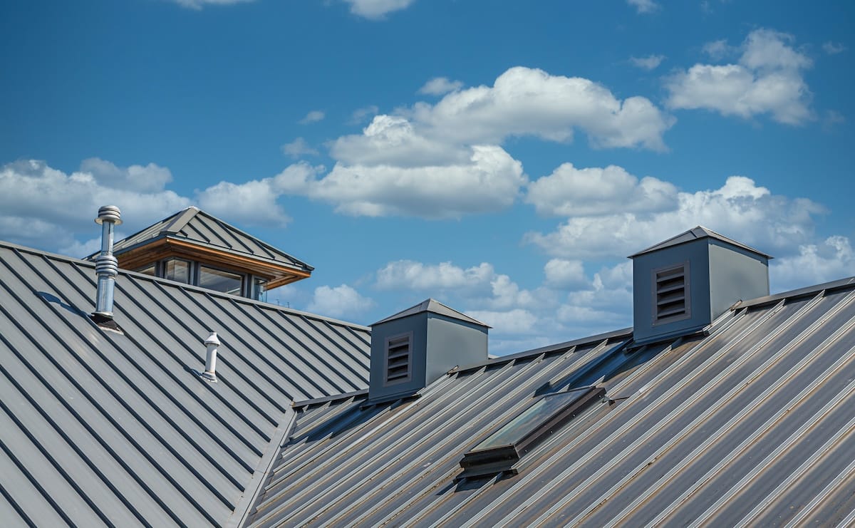 A metal roof with ridges and multiple vents and chimneys against a bright blue sky dotted with fluffy white clouds.