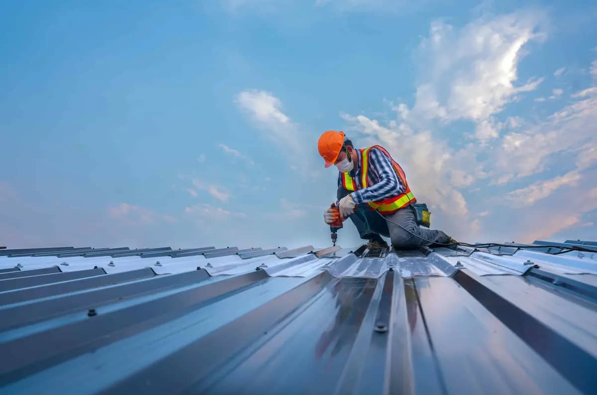 A construction worker wearing an orange helmet and safety vest kneels on a metal roof, using a drill. The sky is blue with scattered clouds in the background.