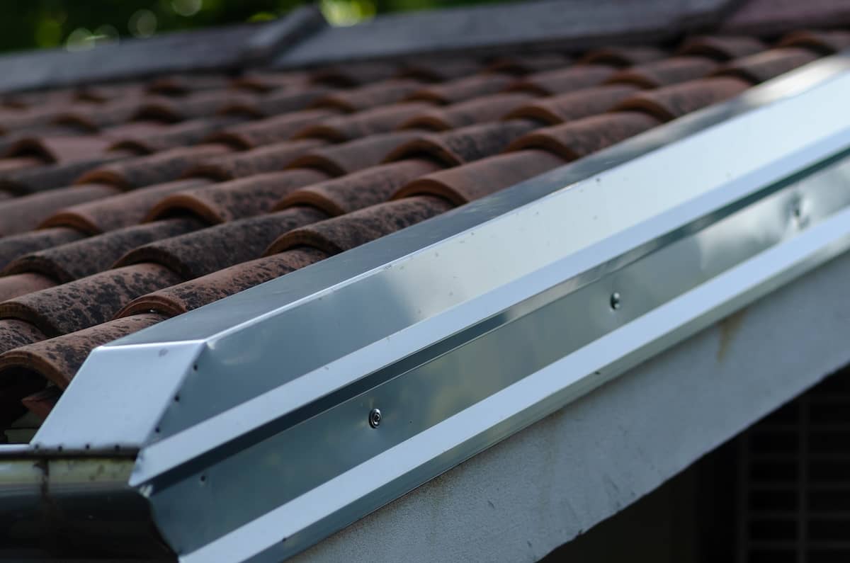 Close-up of a red clay tile roof with a silver metal gutter, showing how to install drip edge along the edge. Screws secure the gutter, and both the roof and gutter appear clean and well-maintained