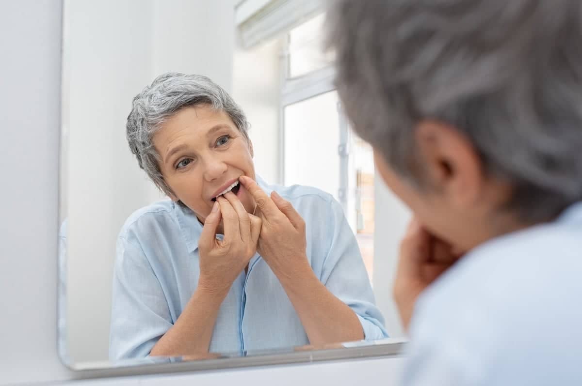 An older adult with short gray hair stands in front of a bathroom mirror, wearing a light blue shirt, and uses floss to clean their teeth. 
