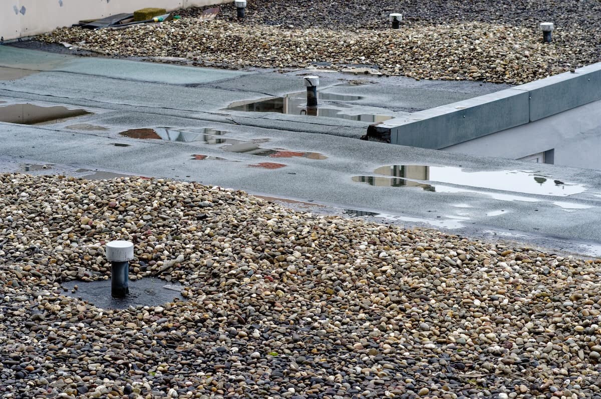 Gravel-covered rooftop with ventilation pipes and patches of standing water reflecting the sky. The surface includes different sections, some with green roofing material, amid scattered pebbles.