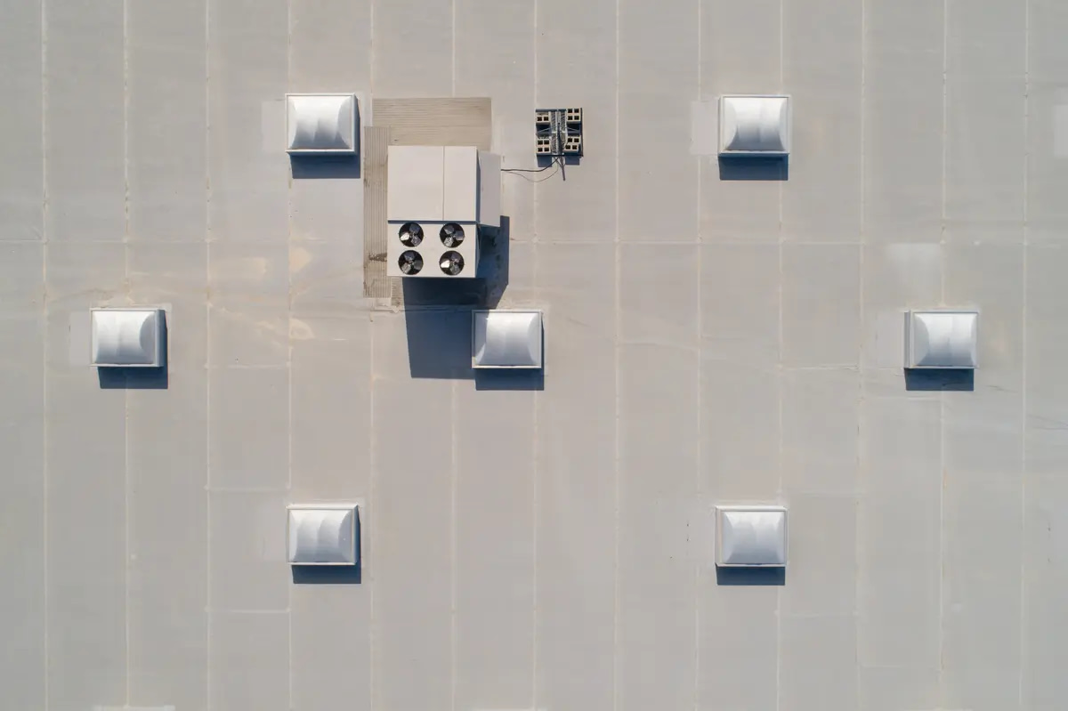 Aerial view of a flat industrial rooftop with a large rectangular HVAC unit and several white square vents evenly distributed across the surface. The rooftop is composed of uniform grey panels.