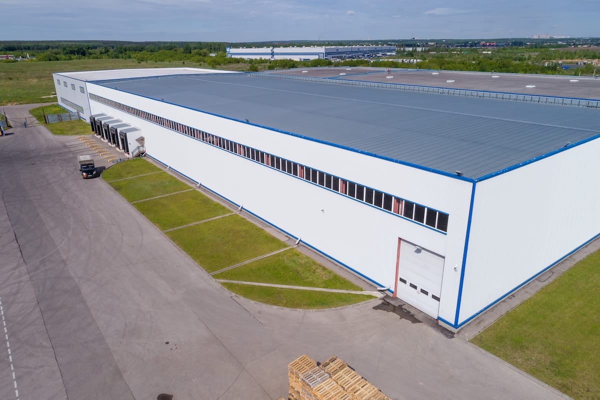 Aerial view of a large, white industrial warehouse with a blue roof and trim, Surrounded by paved roads, grass patches, and some wooden pallets near the building.