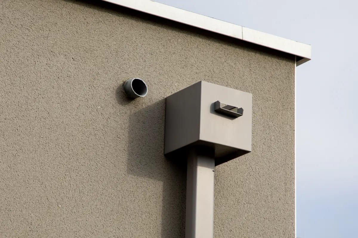 A minimalist architectural detail on a beige building features two protruding elements: a cylindrical vent and a rectangular box with a shadow. The sky is overcast, casting diffused light on the structure.