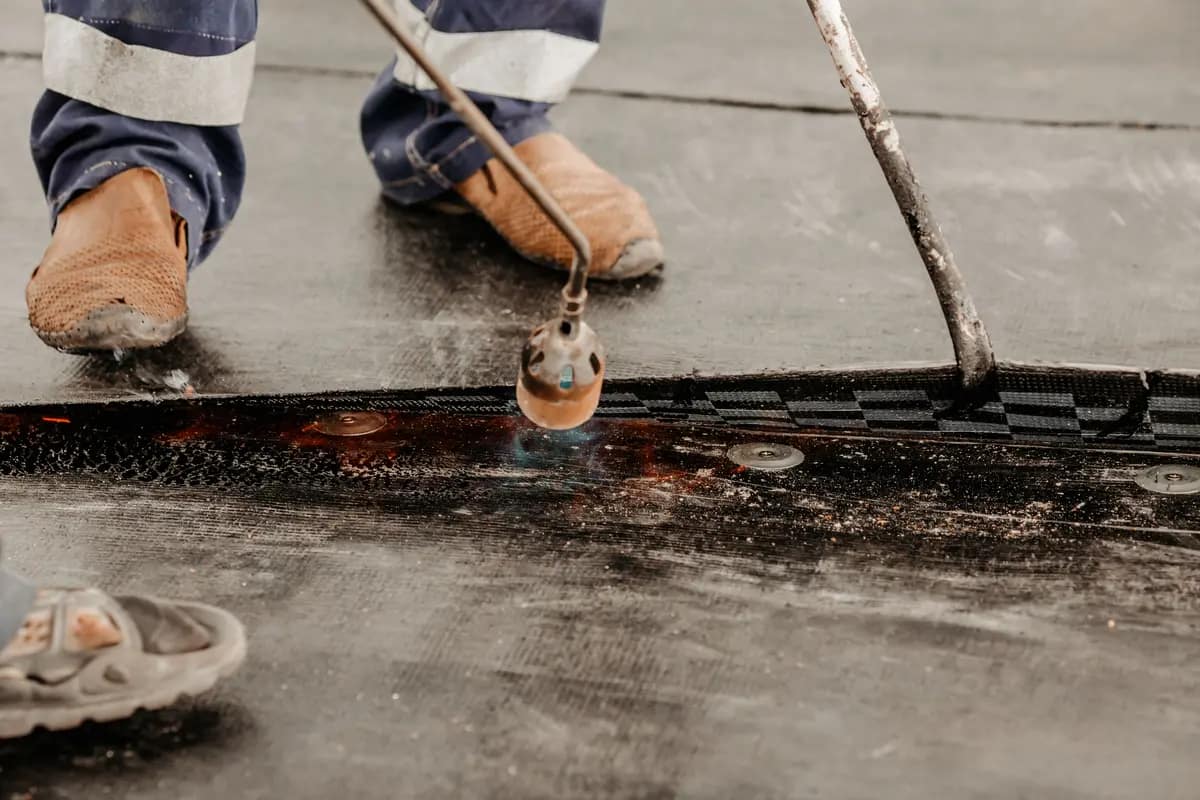 Close-up of a worker using a gas torch to seal a black waterproofing membrane on a roof. The worker is wearing protective boots, and the torch emits a small blue flame onto the membrane.