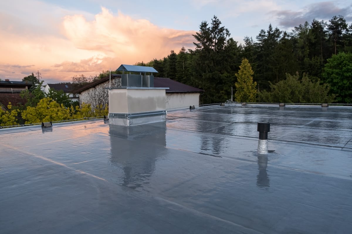 A flat rooftop with a metallic reflective surface, featuring a chimney structure and ventilation pipes. Surrounded by trees and buildings, with a scenic sky displaying soft, colorful clouds at sunset.