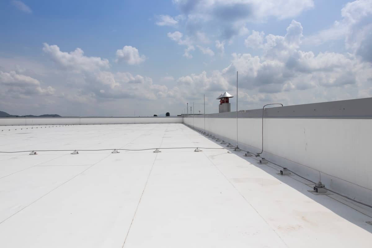 Expansive white flat rooftop with minimal structures, under a partly cloudy sky. An observation tower is visible in the distance along a white fence.