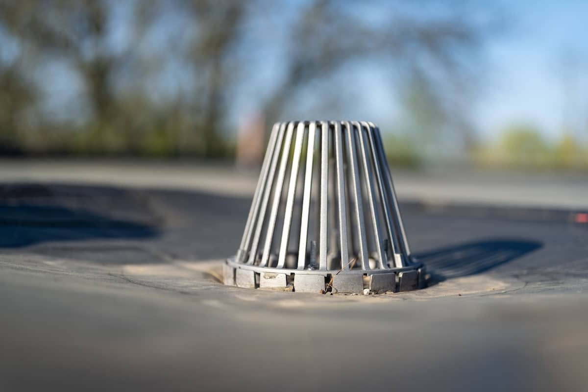 A close-up of a rooftop drain with a metal guard that looks like a cage. The background is blurred, showing hints of trees and clear blue sky.