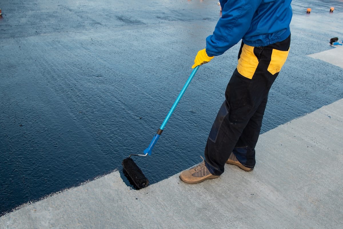 A person using a paint roller to apply a dark, waterproof coating to a concrete surface