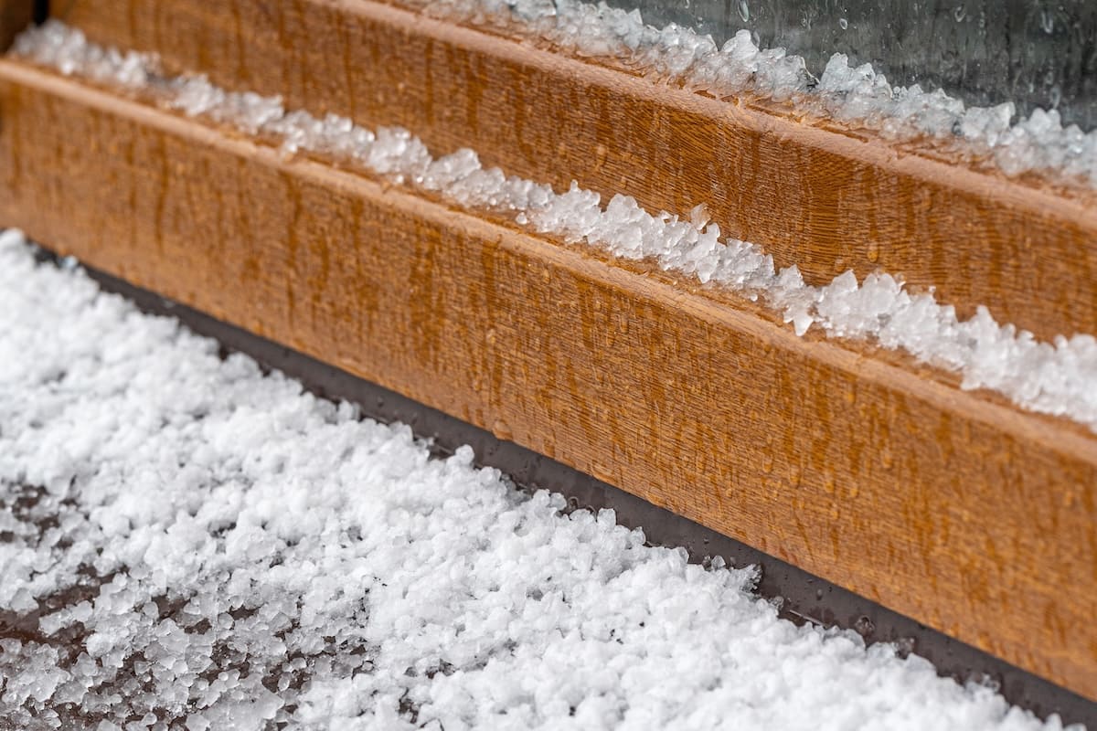 Close-up of a wooden ledge covered with small, white hailstones after a hailstorm, with ice accumulating along the edges—hinting at possible flat roof hail damage