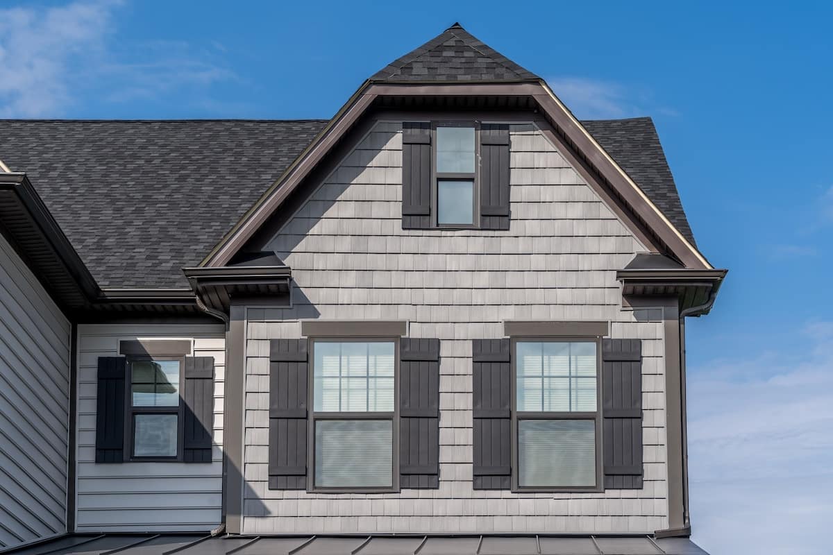 A close-up of a house exterior with light gray siding, black shutters, and three windows beneath a sharply peaked gable roof