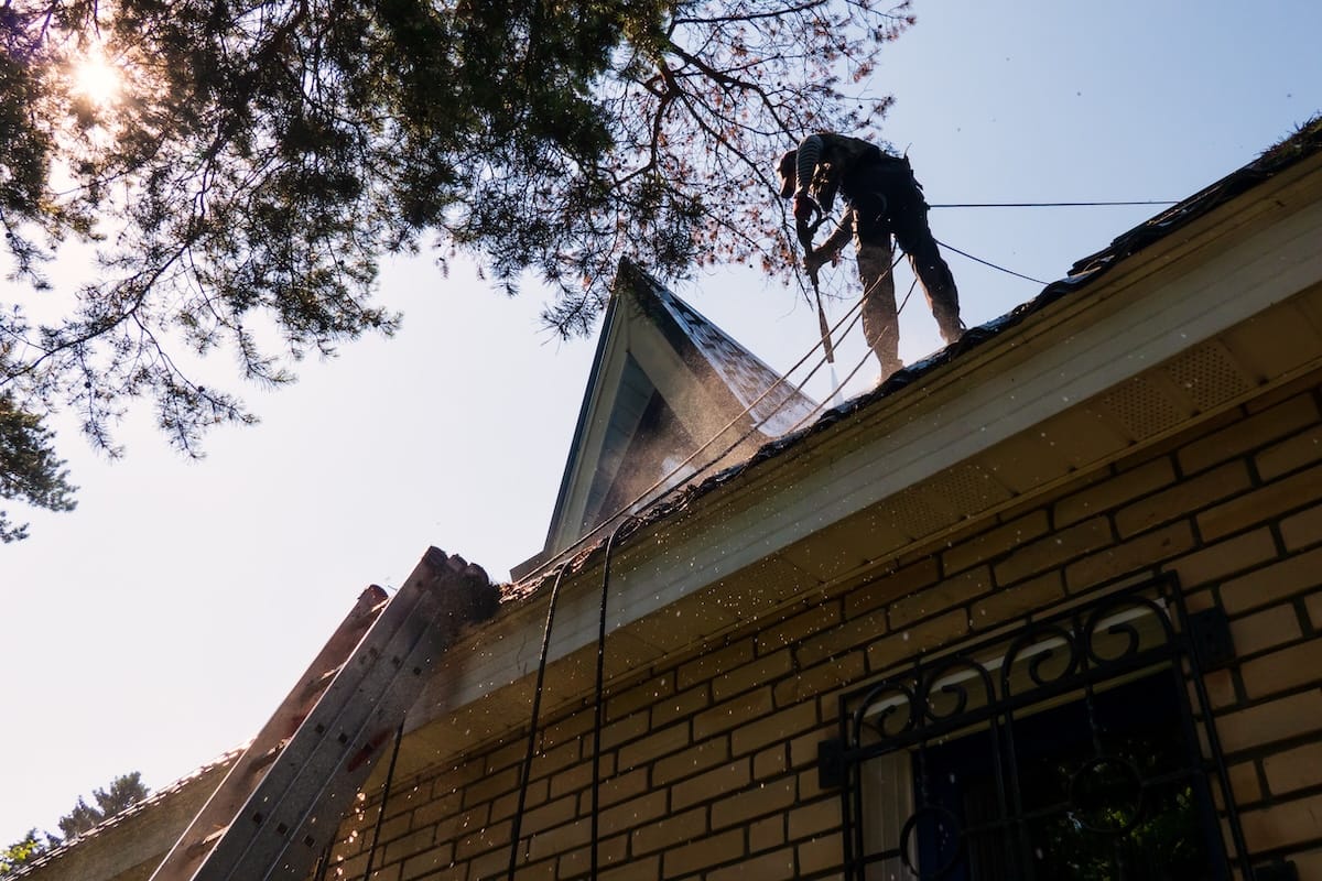 A man stands on the roof of a house and washes metal tiles with a stream of water. Flying drops of water. The bright sun is visible through the branches of the tree.