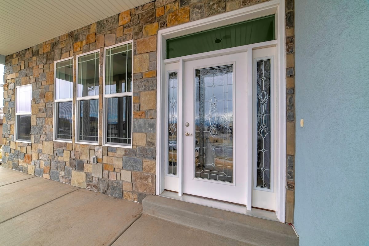Beautiful glass front door with sidelights and transom window at the home facade. Row of window are installed on the stone exterior wall.