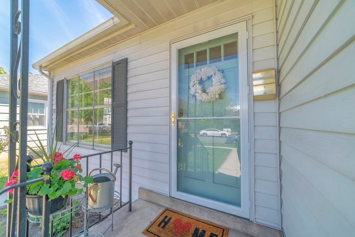Front view of house on real estate listing with glass storm door and green door. Porch with railing, window with shutters, and white wall siding can be seen at the home exterior.