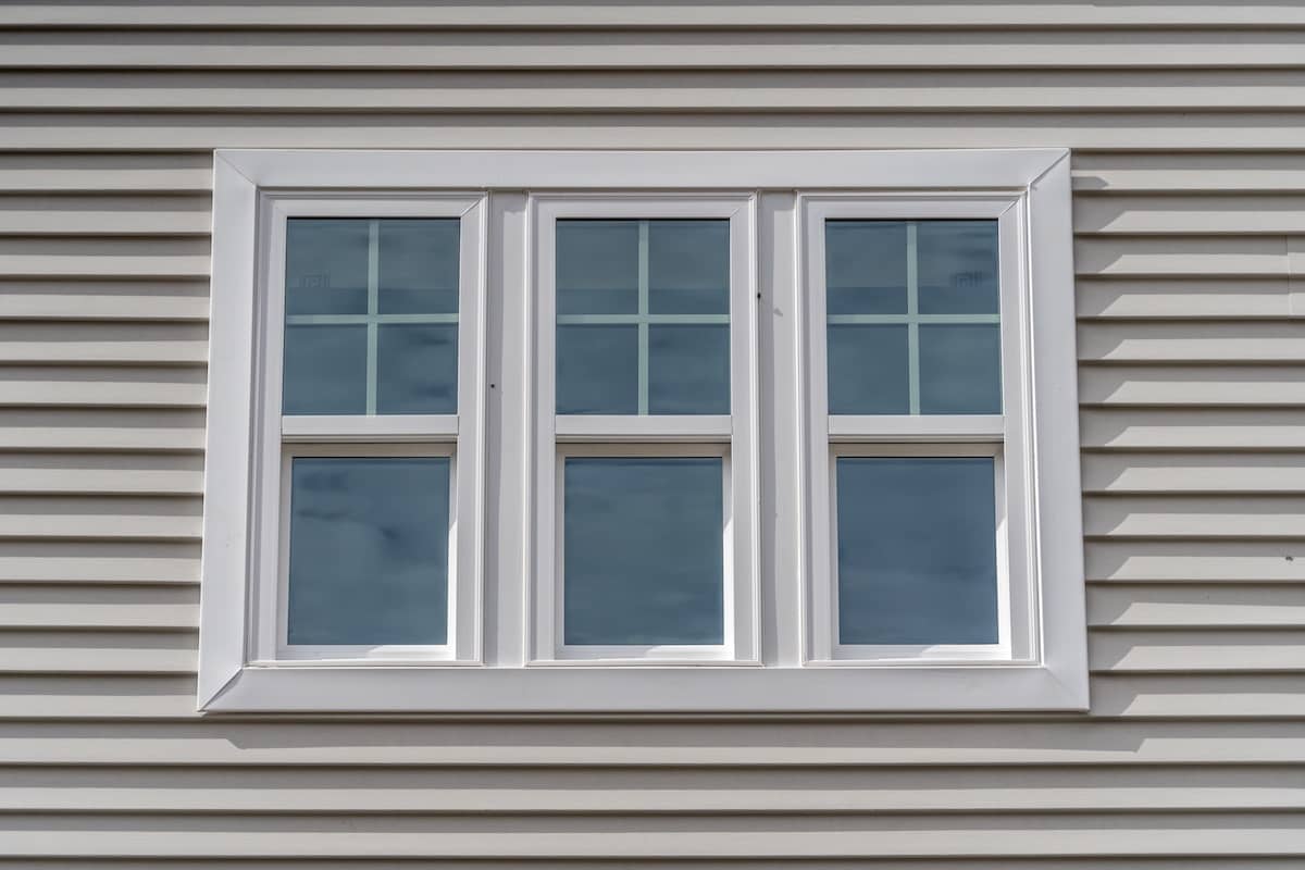 Triple hung vinyl windows with fixed top sash and bottom sash that slides up, sash divided by white grilles a surrounded by white elegant frame horizontal white vinyl siding on a new construction residence