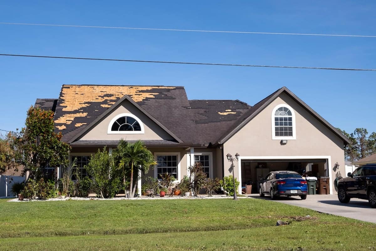 A suburban house shows storm damage with a roof missing shingles. The open garage reveals a blue car inside. Surrounded by a well-kept lawn and various plants, the home awaits roof repair under Durham's clear blue skies.