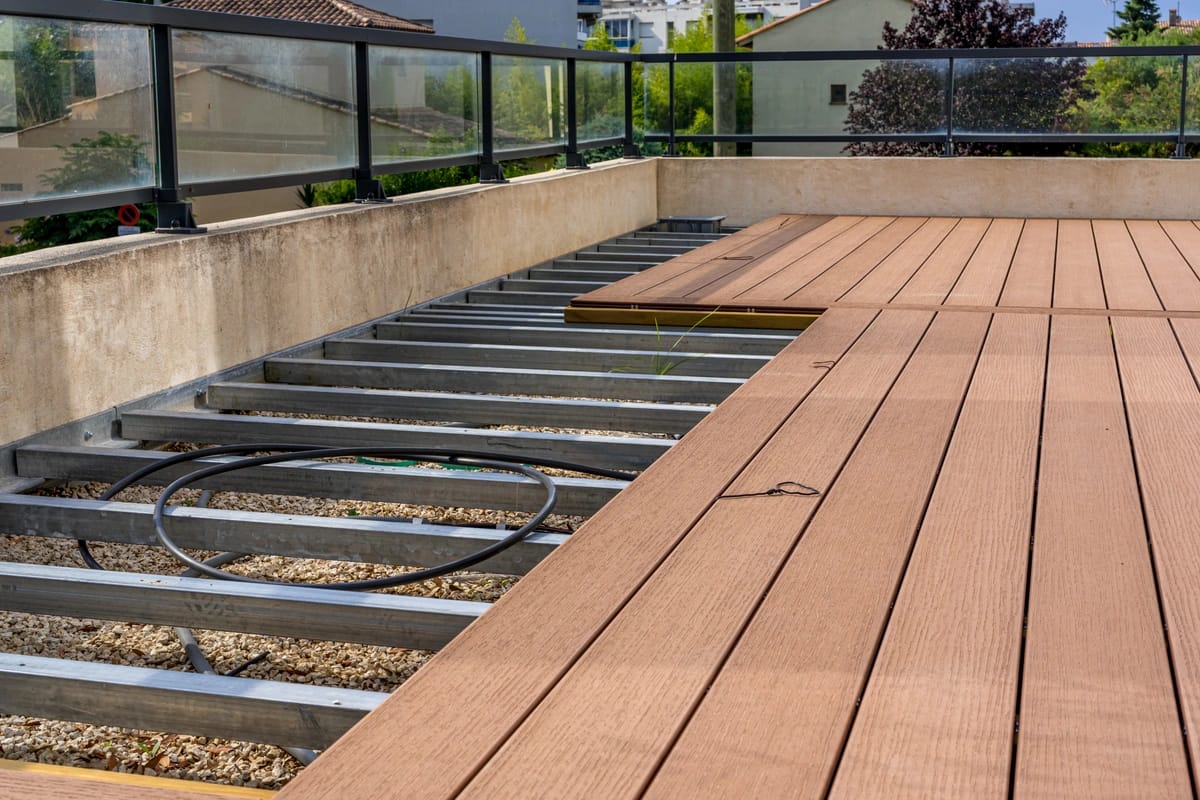 A partially completed wooden terrace with some planks installed on a metal frame hints at the process of how to replace deck boards. An exposed section reveals gravel beneath, while the area is bordered by a glass railing, surrounded by buildings and trees in the background.
